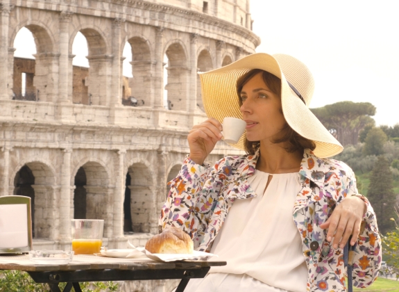 Happy young woman tourist drinking coffee and juice with cornetto at the table outside a bar restaurant in front of the Colosseum in Rome.