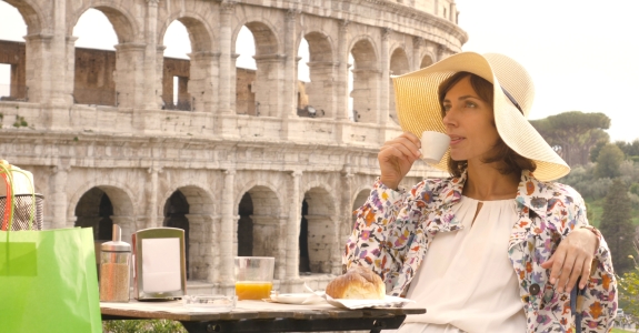 Happy young woman tourist drinking coffee and juice with cornetto at the table outside a bar restaurant in front of the Colosseum in Rome.
