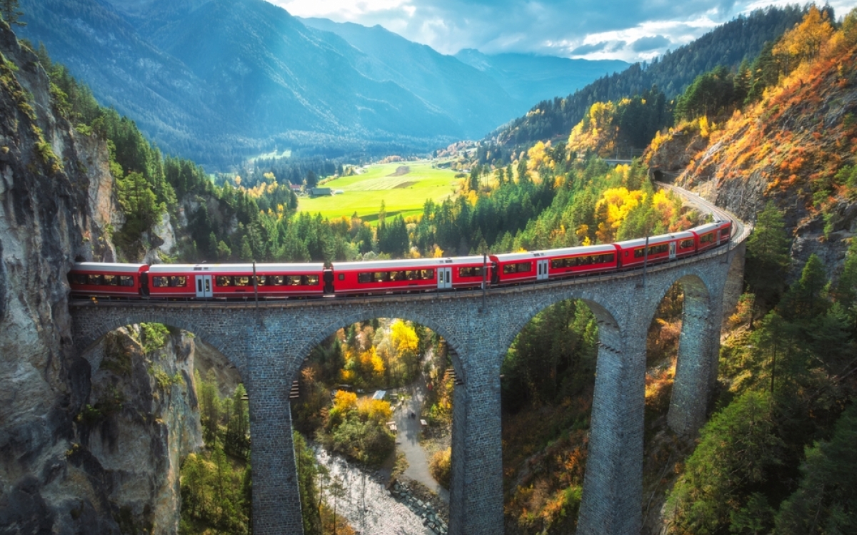 Aerial view of modern red train on Landwasser viaduct in alpine mountains, orange forest at sunset in autumn. Bernina Express, Switzerland. Top view of train in Alps, railroad, colorful trees in fall