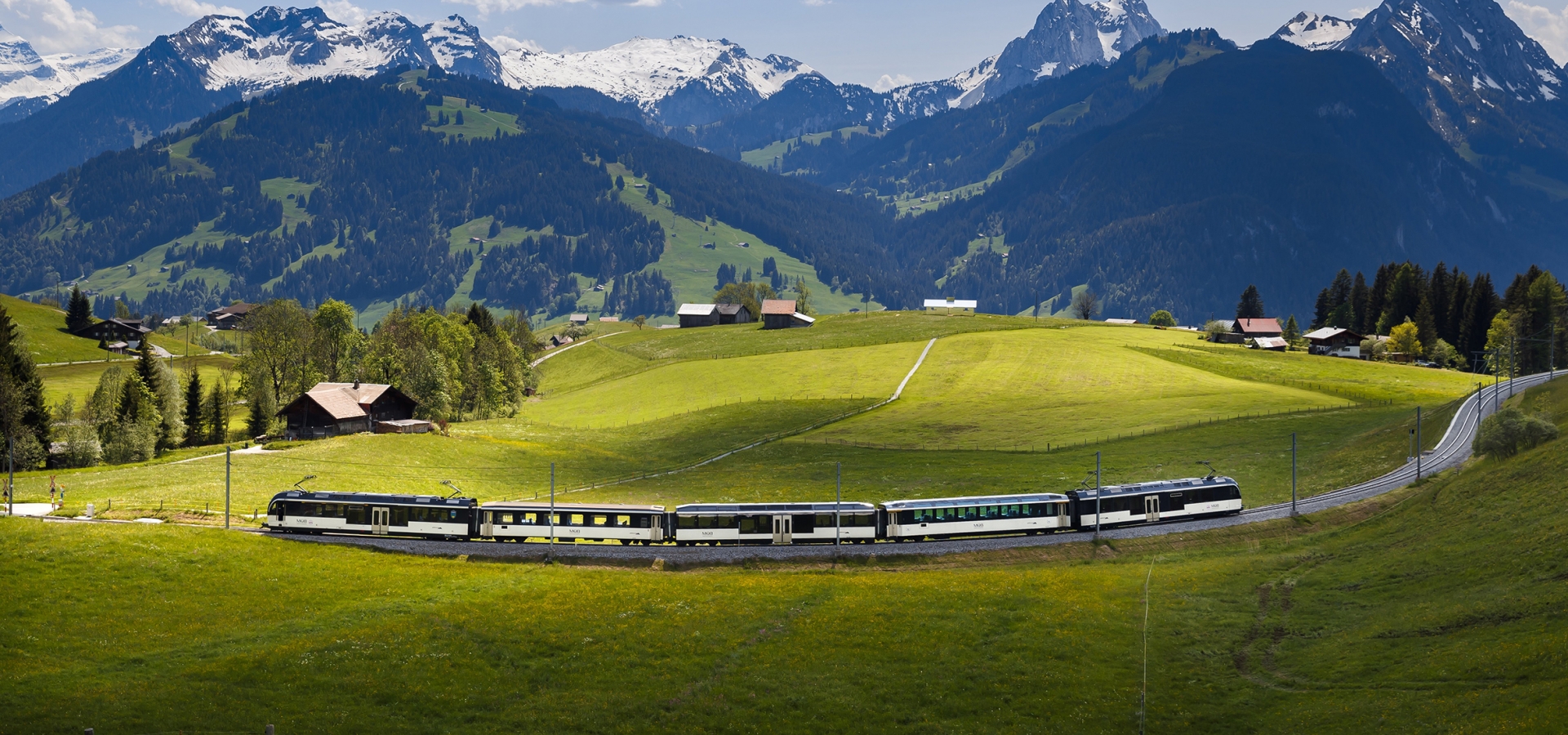 A train travels through a valley with high mountains in the background