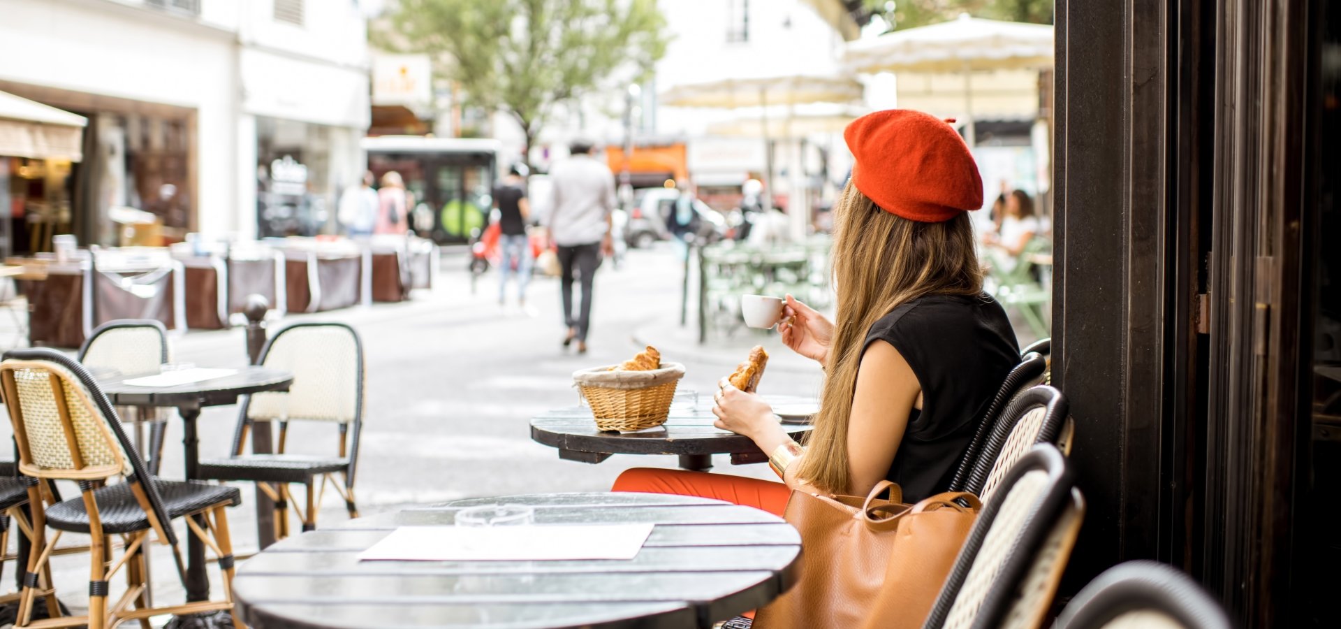Young stylish woman in red beret having a french breakfast with coffee and croissant sitting oudoors at the cafe terrace