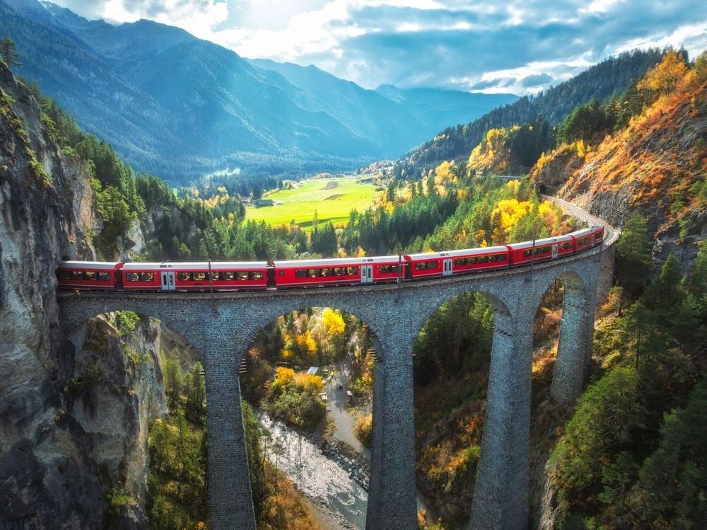 Aerial view of modern red train on Landwasser viaduct in alpine mountains, orange forest at sunset in autumn. Bernina Express, Switzerland. Top view of train in Alps, railroad, colorful trees in fall