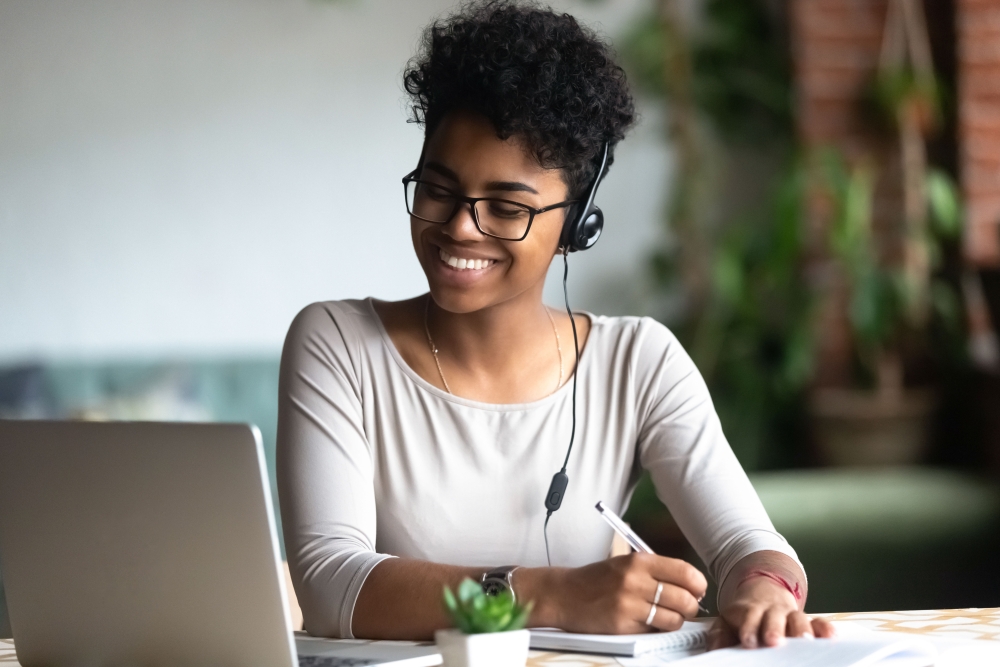 Smiling millennial female student in headphones and glasses sit at desk watch webinar making notes, happy biracial young woman in earphones work study using computer