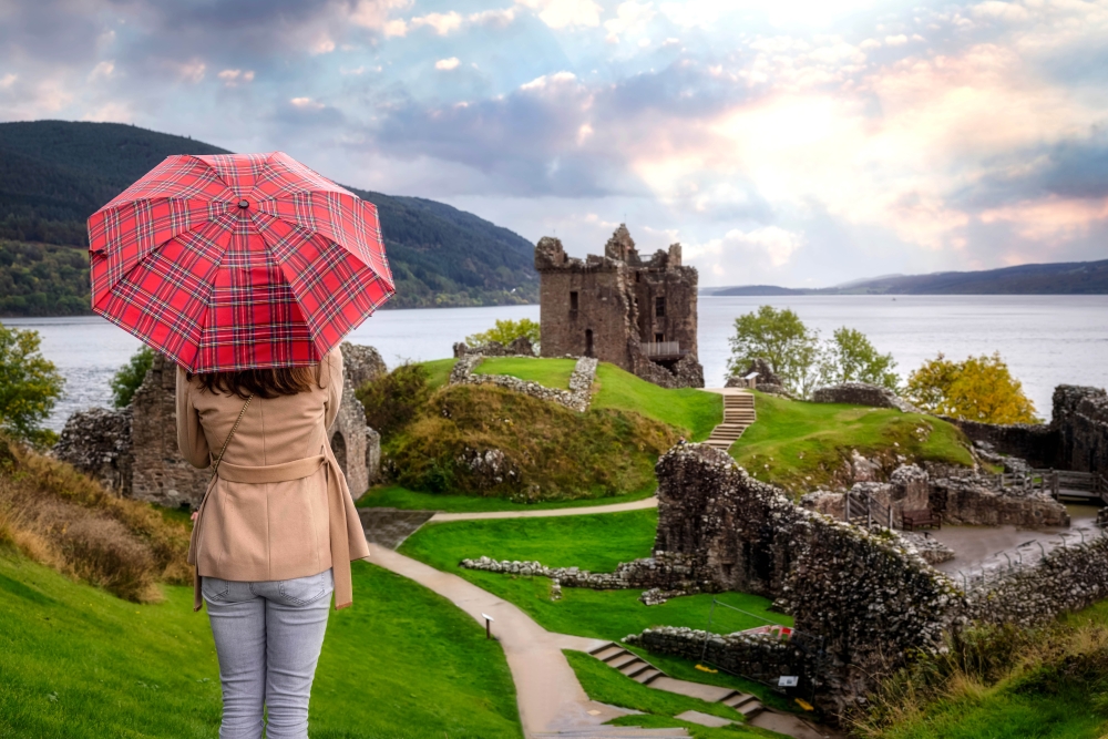 A tourist woman with a scottisch pattern umbrella looks at the famous Urquhart Castle at Loch Ness during autumn time, Scotland